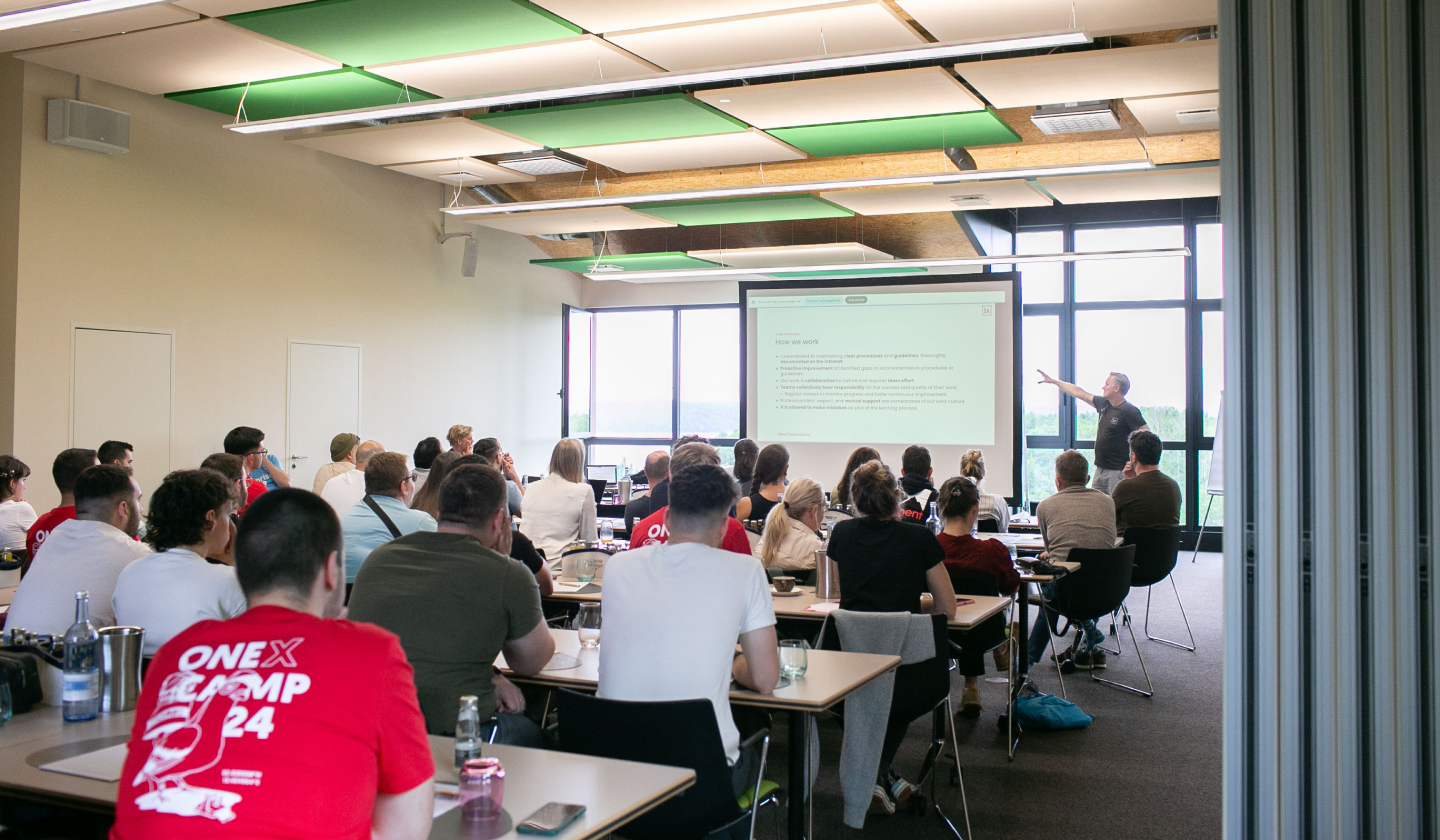 A room full of people watch a person give a presentation in front of a big screen.